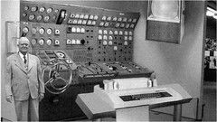 Black-and-white image of a first-generation computer with a large control panel and a man standing beside it.